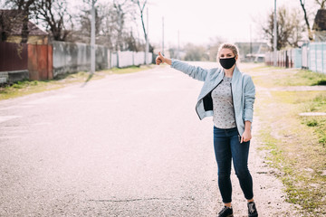 A young woman in a black medical mask in a turquoise jacket and a gray T-shirt stands on the road with a raised hand in the street in the village and catches a car. Quarantine concept. Virus.