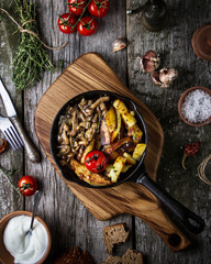 Fried potatoes with mushrooms mushrooms in a pan with tomato on a wooden board on an old rustic table, top view.