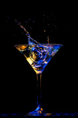 Copa de martini salpicando con líquido en tonos dorados, azules y púrpura, sobre fondo negro.