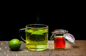 Taza de vidrio con té de limón, un limón entero y frasco con miel, sobre mantel con fondo negro