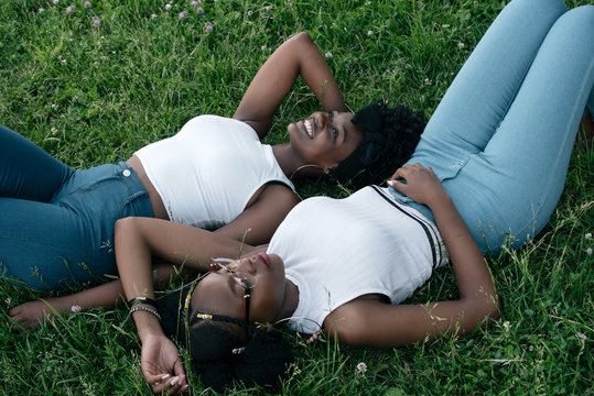 Two african girls in jeans and white tops are laying on the grass and smiles