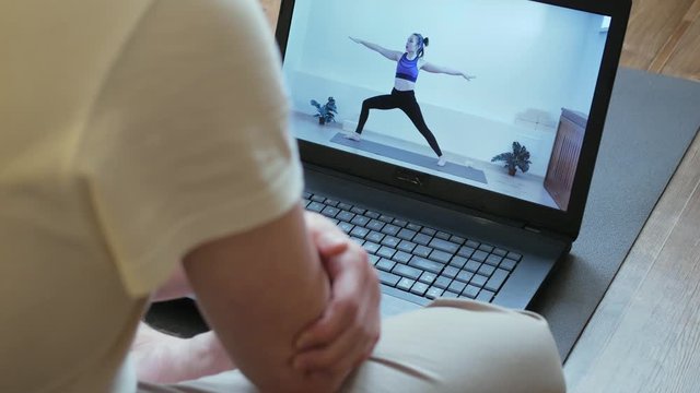 A man sits on a yoga mat at home on self-isolation, watches yoga classes online using a laptop, a girl trainer shows virabhadrasana.