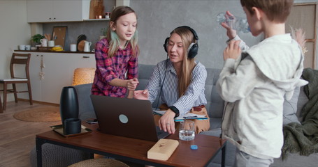 Beautiful Caucasian mother trying to work home online using laptop, distracted by two teenage...