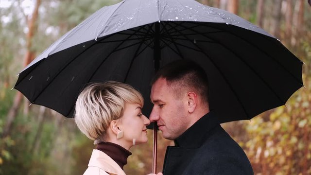 Woman and man stand under black umbrella in forest. People stand close to each other. Couple spends time outside together.