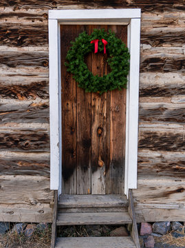 Green Christmas Holiday Wreath with Red Bow Hanging On Knotted Wood Grain Door Inset Into Old Log Cabin