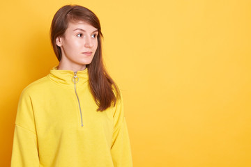 Image of charming woman with perfect skin and pleasant appearance, posing isolated over yellow studio background and looking aside, lady wearing hoodie. Copy space for advertisement or promotion.