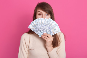 Indoor shot of astonished woman standing against pink studio wall, covering her face with fan of money, posing with big eyes, woman wearing casual shirt, wants to spend money for new clothing.