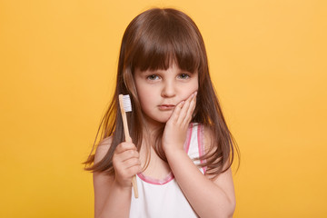 Closeup portrait of serious cute child keeping palm on cheek, having toothache, holding toothbrush...
