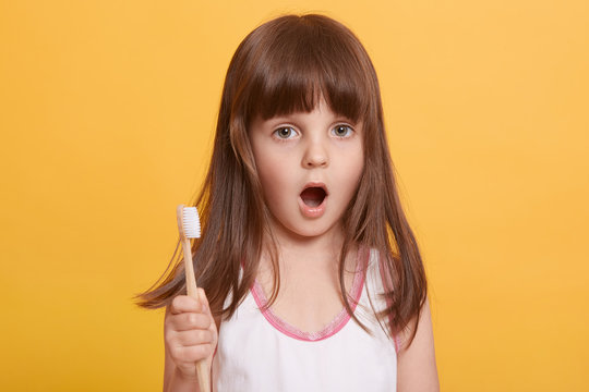 Picture of astonished little girl with opened mouth and holding tooth brush in hand, looking directly at camera, suffering from toothache, child's first teeth falling. Dental hygiene concept.