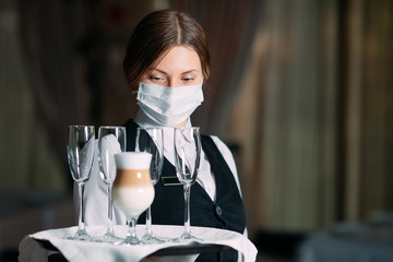 A female Waiter of European appearance in a medical mask serves Latte coffee.