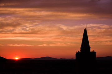 Obraz premium Sun hiding on the horizon, backlit a tower with the silhouettes of various storks
