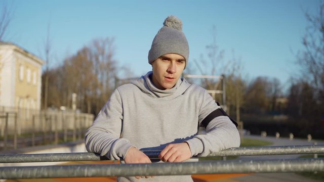Panning medium shot portrait of athletic young man looking at camera while recovering after exercise leaning on parallel bars at street workout spot