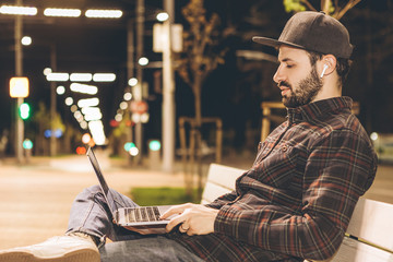 stylish bearded young man dressed in casual clothes works with a laptop in his hands sitting in the evening on a city street