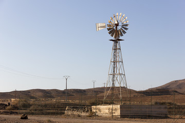 Rusty metal windmill in rural area at sunset. Agrarian old building structure and electric posts on background in Fuerteventura island, Spain. Remote location, desert, nobody concepts