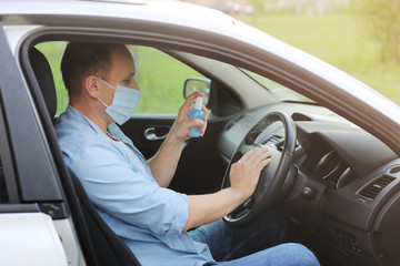 Spraying antibacterial sanitizer spray on steering wheel car, infection control concept. Prevent Coronavirus, COVID-19, flu. Man wearing in medical protective mask driving a car. Disinfecting wipes.