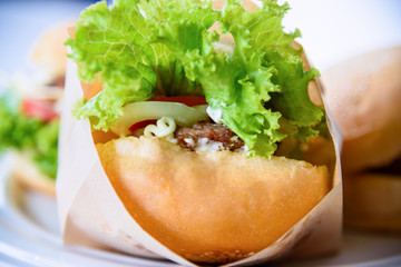 Burger is delicious American fast food, Closeup a hamburger made from pork or beef, green lettuce bread onion and tomato in a paper bag and plate on table background at home to picnic or party