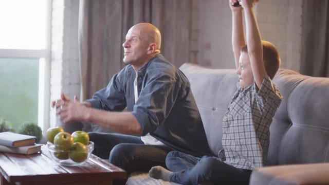Handheld shot of happy father and son smiling and playing video game at home, then celebrating after winning match
