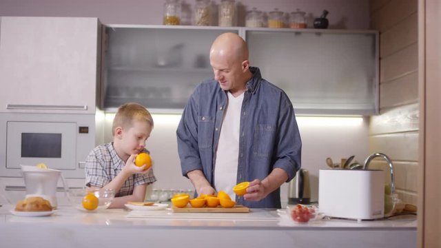 Tracking Of Bald Man In Jeans Shirt Talking To Cute 8 Year Old Son And Cutting Orange In Half In Kitchen