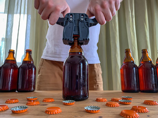Craft beer brewing at home, man closes brown glass beer bottles with plastic capper on wooden table with orange crown caps. - obrazy, fototapety, plakaty