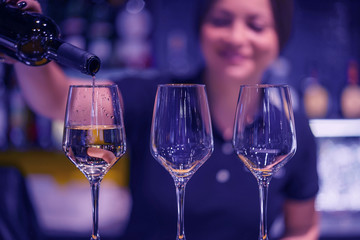 Bartender woman smiling, pours white wine into a glass from a bottle. Night time.