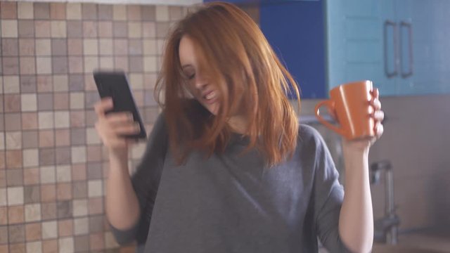 Girl with red hair in morning in kitchen listens to music, sings and dances. Young redhead woman dancing with a cup of coffee and a phone in her hands. Happy hipster millennial girl in t-shirt.