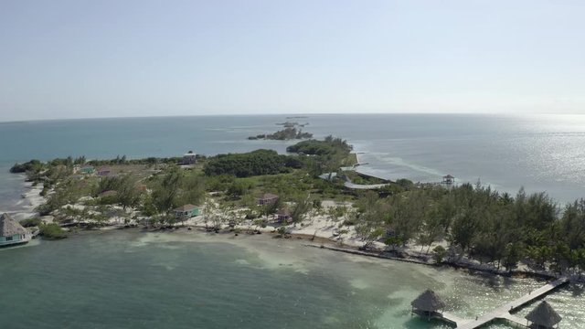 Aerial panning shot of trees and resorts on island amidst sea, drone flying over beach during sunny day - Coco Plum Cay, Belize