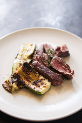 Meat fillet with grilled zucchini on a white dish with a white knife, top view over a black rustic board