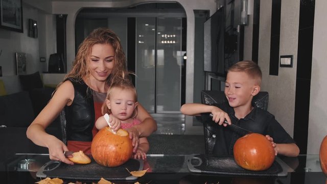 Mother is in kitchen with two kids. Boy and little girl learn how to cut pumpkins. Family prepares decorations for halloween.