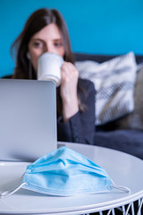 Protective mask for coronavirus on a table and business woman working from home and drinking a coffee in the background. Covid-19.