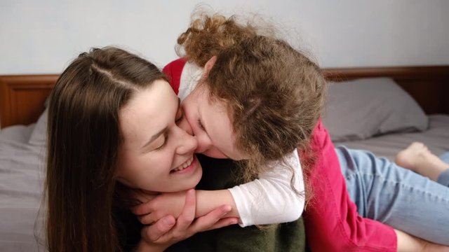 Cute adorable funny kid daughter kissing on cheek young mum  playing on bed at home, smiling mother or nanny rest take, little girl enjoy tender moment with mom, love. Relative people family concept