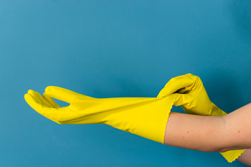 Close up on hand of unknown caucasian woman female girl putting on adjusting holding protective yellow rubber gloves to clean or disinfection against blue wall background in day