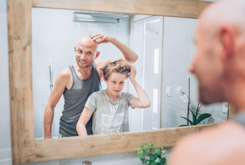 Bald dad and his long-haired teenager son in bathroom in front of the mirror. Father showing to boy his new style haircut. Common family lifestyle and parenting concept.