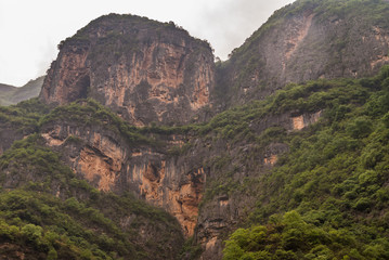 Wuchan, China - May 7, 2010: Dawu or Misty Gorge on Daning River. Tall huge black and brown cliffs with green foliage in spots under silver sky.