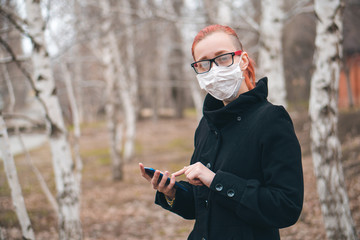 Young beautiful woman in medical mask holds phone in hand in park and looks at camera. Health care or epidemic concept with copy space.