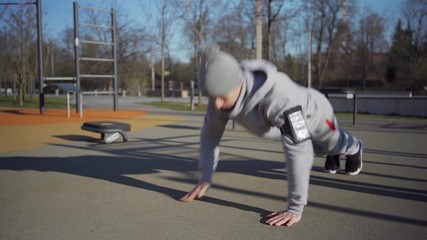 Athletic young man doing variations of shoulder tap push-ups at street workout spot outdoors, armband with cellphone on his arm