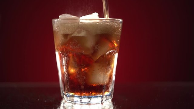 A transparent glass with a bunch of ice is filled with Coca-Cola. A glass stands on a wet table with water droplets on a red background. Soft drink soda