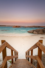Wooden staircase leading down to a beach at dusk with orange granite rocks in the water and soft color in the sky caused by bush fire smoke haze at Twilight Cove, Esperance, Western Australia
