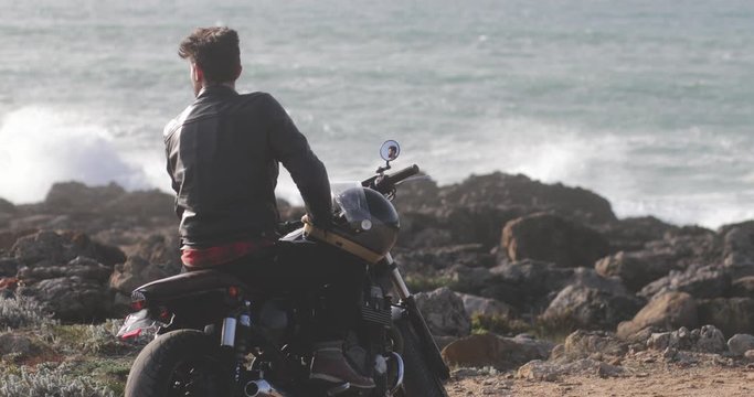 Back view man sitting on classic motorcycle gazing at seafront