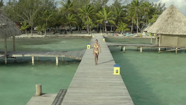 Aerial: Drone shot of young woman in bikini walking on pier over sea during sunny day, sensuous female is on vacation - Coco Plum Cay, Belize