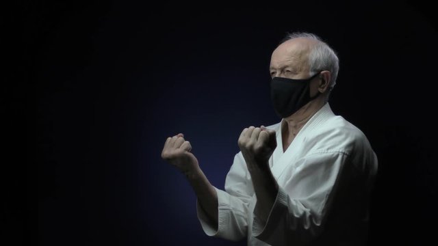In a black medical mask, an old athlete is training blocks against a dark background