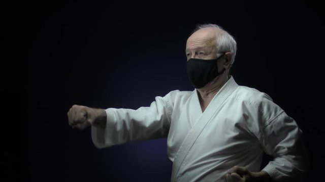 In a black medical mask, an old athlete makes blocks and punches against a dark background