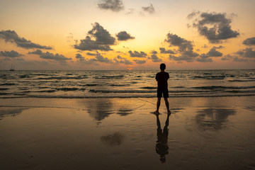 Fototapeta premium The silhouette of a man by the sea at sunset, at the beach and beautiful sky at Ko Kood, Thailand, the black shadow of a person and the clouds reflecting the water in warm orange tones.