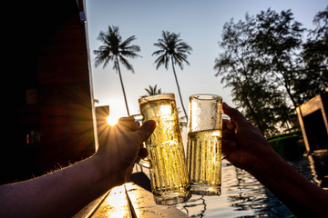 The hand of two people holding a glass of beverage and cheer each other to celebrate in the pool In the tropical garden atmosphere, there are coconut trees and sun rays. Concept of celebrating success