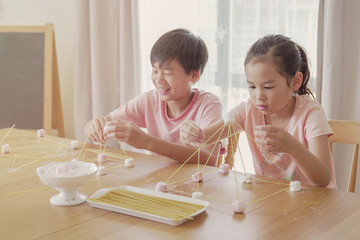 Mixed race young Asian children building tower with spaghetti and marshmallow learning remotely at...