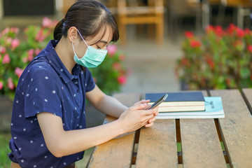 Young Asian student girl wearing medical mask and holding smartphone, looking at screen, using app or messaging while sitting at garden bench with book. new normal concept after covid-19