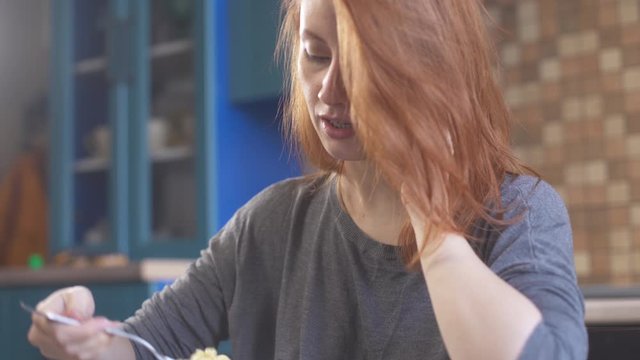 Hungry girl with red hair eating a noodles and talking on the phone. Уoung redhead woman enjoying a food. Hipster girl in kitchen eating chinese food. Delicious fast food. Healthy nutrition. Close up.