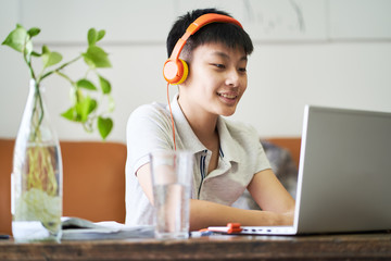 Asian teenage boy studying at home during pandemic wearing headset and smiling