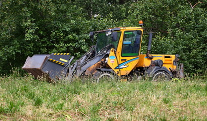 A small yellow the tractor in green grass on a background of trees