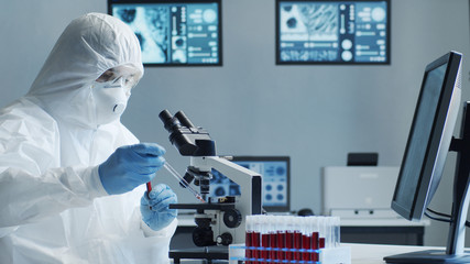 Scientist in protection suit and masks working in research lab using laboratory equipment: microscopes, test tubes. Coronavirus 2019-ncov hazard, pharmaceutical discovery, bacteriology and virology.