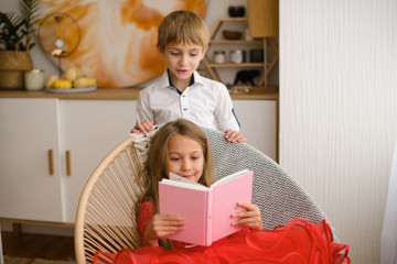 girl with her hair in red dress is reading pink book sitting in wicker chair. Her brother looks over her shoulder.